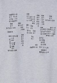 T-shirt grigia chiara con motivi testuali neri distribuiti. Il testo include "Obey Studios" in diverse orientamenti e dimensioni, con un effetto usurato.