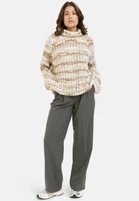 Fleckiger Strickpullover in Creme- und Braunstreifen, hoher Kragen, kombiniert mit maßgeschneiderten grauen Hosen und weißen Sneakers mit metallischen Akzenten.