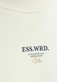 Krems krāsas audums ar ribotām apkaklēm; tajā ir tumši zilā un zelta burti "ESS.WRD." un "Kopš 1990. gada Essentials Dept."