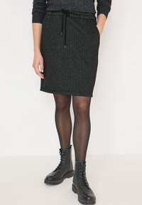 Vrouw in een donkergrijze herringbone rok, zwarte doorzichtige panty en zwarte veterschoenen, staand met één hand in haar zak.