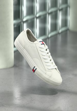 White Tommy Hilfiger sneaker with laces floating above a gray surface, glass block wall and column in soft-focus background.