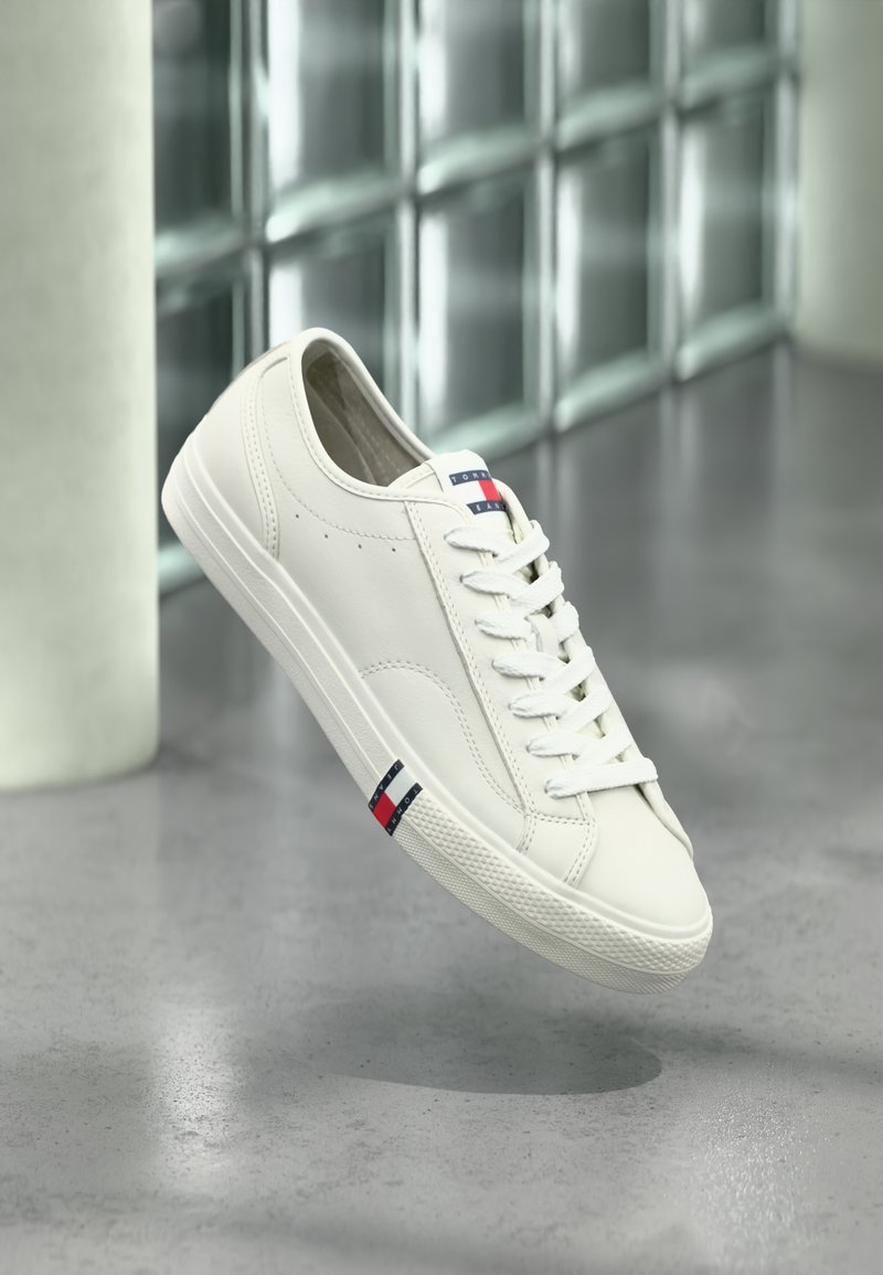 Baskets blanches Tommy Hilfiger avec lacets flottant au-dessus d'une surface grise, mur en verre et colonne flous en arrière-plan.