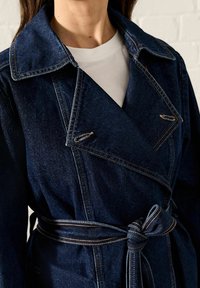 Primer plano de una persona que lleva un abrigo trench de denim azul oscuro con solapas anchas y un cinturón atado sobre una camiseta de cuello redondo blanca.