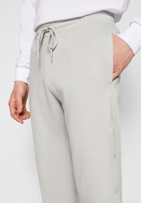 Light gray sweatpants with an elastic waistband, drawstring, and side pockets. Features snap button detail along the legs for versatile styling.