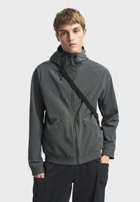 Grå, letvægtsjakke med hætte, lynlåslukning foran og elastiske manchetter. Har vinklede sømdetaljer og sidelommer.