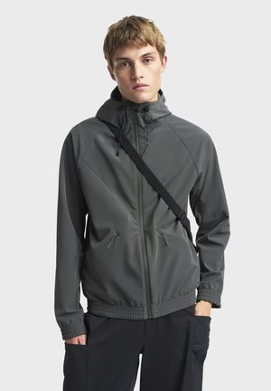 Chaqueta gris, ligera, con capucha, cierre frontal con cremallera y puños elásticos. Presenta detalles de costura angulares y bolsillos laterales.