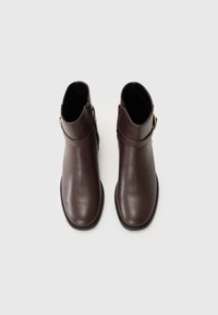 Botas de tobillo de cuero marrón con una textura suave, cremallera lateral y un acento decorativo de hebilla. Suela negra plana, diseño minimalista.