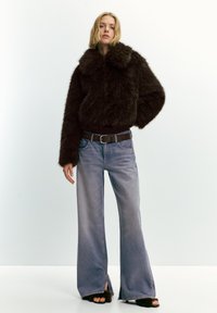 Žena v tmavě hnědém kožichu, s širokými vybledlými modrými džíny s předními rozparky, hnědým páskem a černými koženými sandály stojící na jednobarevném pozadí.