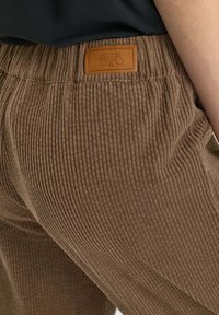 Primer plano de unos pantalones de pana marrones con cintura elástica y etiqueta de cuero color marrón claro en la parte posterior, combinados con una blusa negra.
