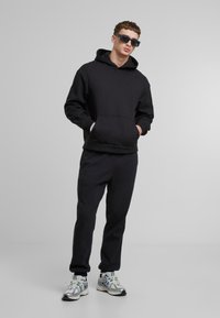 Svart hettegenser med kengurulomme, parret med matchende svarte joggebukser og metallisk sølvfargede joggesko. Modellen bruker mørke solbriller.