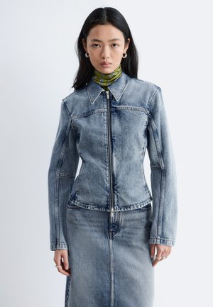 Kobieta ubrana w jasnoniebieską kurtkę dżinsową na zamek i dopasowaną dżinsową spódnicę, pod spodem zielony top z wysokim kołnierzem w wzory, stojąca na białym tle.