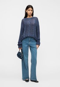Pull en maille bleu marine avec tissage ouvert, manches larges, associé à un jean taille haute bleu clair. Accessoires comprenant un petit sac à main à motifs.
