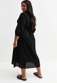 Black linen midi dress with 3/4 sleeves and embroidered accents. Features a tie waist and relaxed fit, complemented by flat sandals.