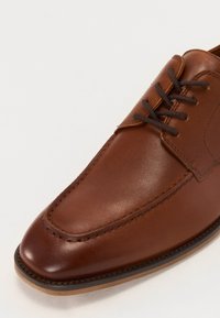 Brune lærsko med rund tå, syet detaljering og brune snørebånd. Har en glat tekstur med en let blank finish.