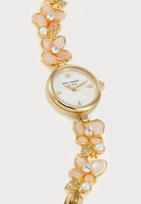 Reloj de tono dorado con diseño de pulsera floral, que cuenta con flores de esmalte rosa, cristales transparentes y perlas. Esfera blanca con logo y acentos dorados.