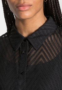 Sort bluse med et struktureret chevron-mønster, med knaplukning foran og en klassisk krave. Lavet af et gennemsigtigt materiale med en blød overflade.