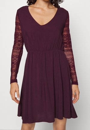 Kobieta ubrana w sięgającą kolan sukienkę w głębokim odcieniu fioletu z dekoltem w serek i długimi koronkowymi rękawami, stojąca na jednolitym jasnym tle.
