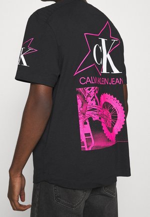 Černé bavlněné tričko s velkým růžovým hvězdicovým grafikou, na které je nápis "CK" a "CALVIN KLEIN JEANS", doplněný ilustrací motocyklu.