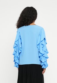 Haut bleu avec des manches longues à volants, texture lisse, encolure ronde, associé à une jupe plissée noire, vu de derrière.
