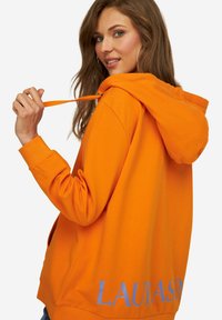 Oranje hoodie met een trekkoord capuchon, ribgebreide manchetten en een groot gedrukt "LAUNASØ" logo in het blauw op de achterkant. Zachte stof textuur.
