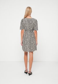 Γυναίκα που στέκεται με την πλάτη προς τα έξω, φορώντας ένα λευκό φόρεμα μέχρι το γόνατο με μαύρες βούλες και μαύρες ψηλές σάντες, σε λευκό φόντο.