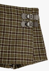 Brun og grønternet kort beklædning lavet af struktureret stof, med to bæltespænder i mørkt læder ved taljen.