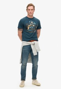 Camiseta de algodón color azul verdoso con un estampado gráfico; sudadera gris claro anudada en la cintura; jeans azules; zapatillas beige; tatuajes visibles en el brazo.