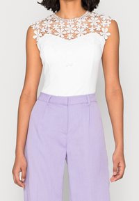 Vit bodysuit med en blommig spetskrage och korta ärmar; stylad med lavendelfärgade högmidjade byxor med en slät textur.