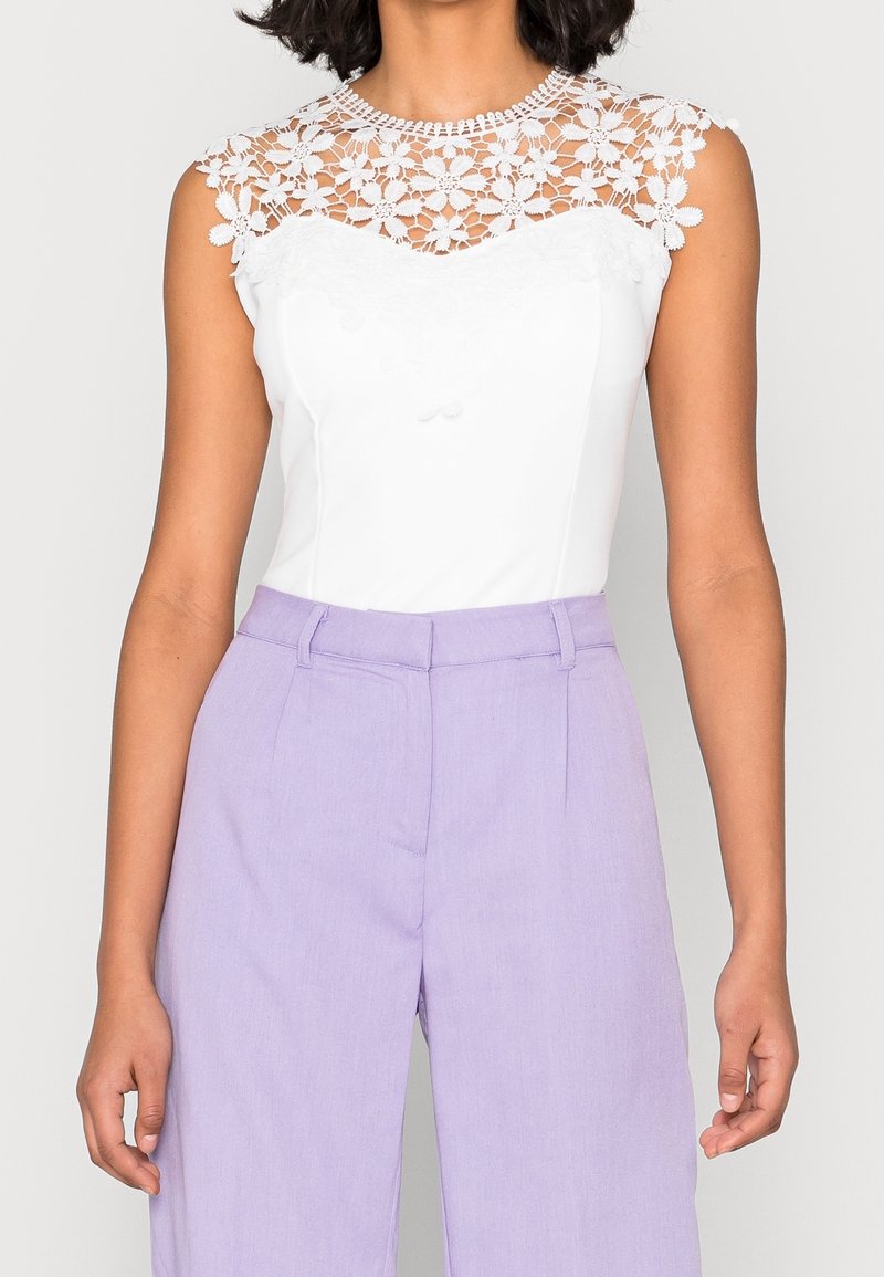 Vit bodysuit med en blommig spetskrage och korta ärmar; stylad med lavendelfärgade högmidjade byxor med en slät textur.