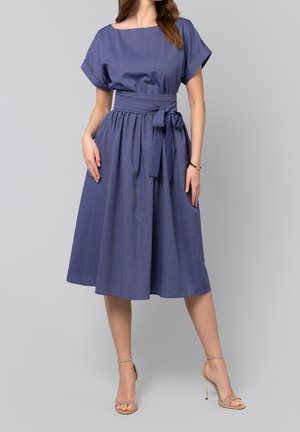 Mujer con vestido azul de manga corta, hasta la rodilla, con cinturón atado en la cintura y sandalias de tacón beige sobre un fondo gris.