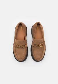 Paire de mocassins en daim marron avec semelles noires et détail en métal doré en forme de mors sur fond blanc.