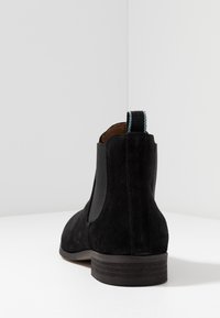 Bottine Chelsea en suède noir avec des panneaux latéraux élastiques, bout arrondi et un petit talon empilé. Le dos présente une languette de tirage accentuée de bleu.