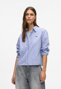 Blå og hvit rutete skjorte med rullede ermer, med en liten brodert logo, lagt over en gråstripet skjorte.