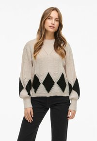 Pletený svetr ve světlé béžové barvě s černým diamantovým vzorem na rukávech, s volným střihem a vroubkovanými manžetami. Měkká textura a složitý design.