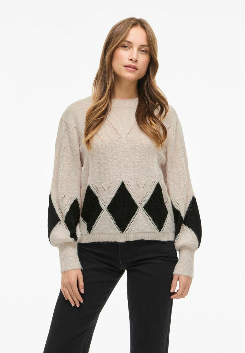 Pletený svetr ve světlé béžové barvě s černým diamantovým vzorem na rukávech, s volným střihem a vroubkovanými manžetami. Měkká textura a složitý design.