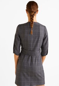 Robe à carreaux gris avec manches trois-quarts, taille cintrée et ceinture. Le tissu semble texturé, avec une silhouette structurée et des détails de couture.