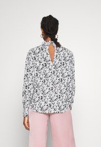 Blusa bianca con motivo floreale nero, design con apertura a chiave sul retro, maniche lunghe e colletto alla cinese. Abbinata a pantaloni rosa a gamba larga.