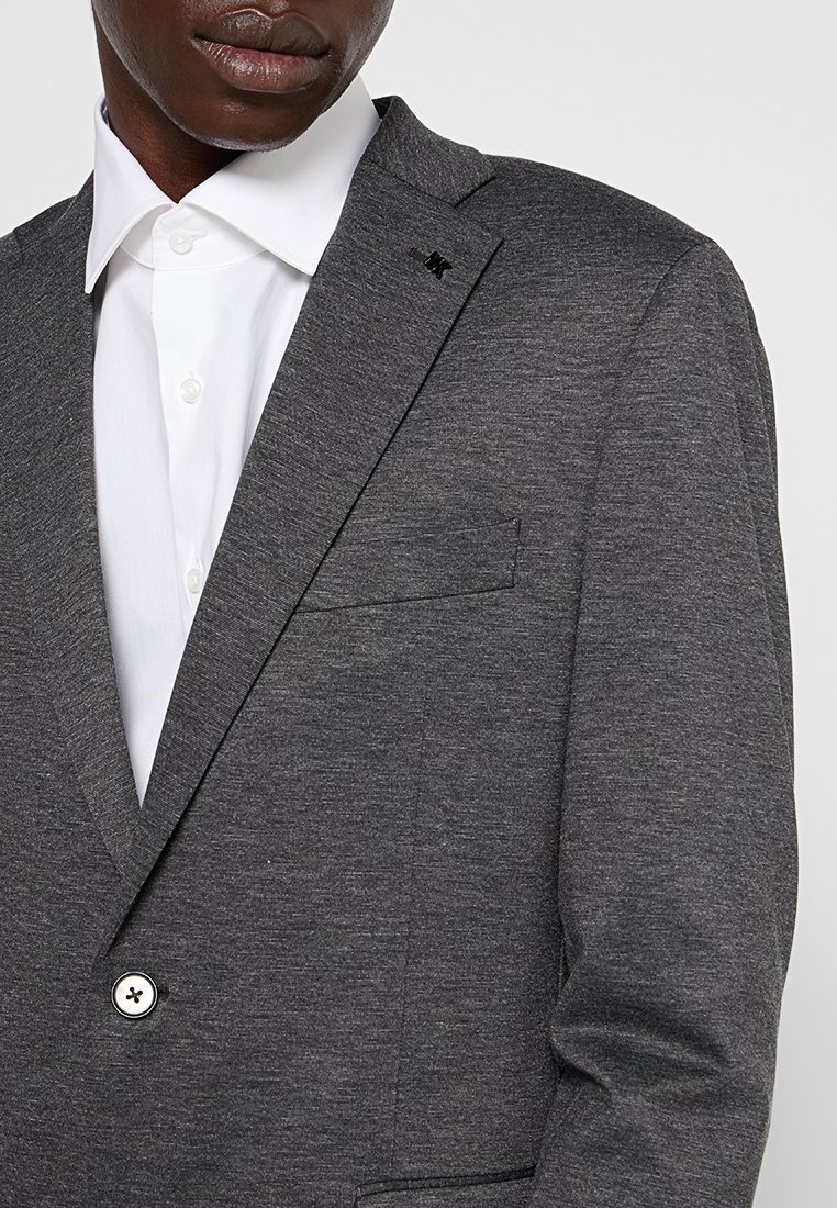 Blazer gris con una tela texturizada, cierre de un solo botón y un pequeño logo en la solapa. Llevado sobre una camisa de vestir blanca.