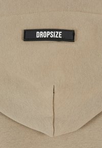 Îmbrăcăminte din țesătură bej cu un buzunar cusut și o etichetă neagră pe care scrie "DROPSIZE" în text alb, având o textură netedă.