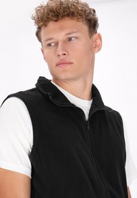 Schwarze Fleece-Weste mit Reißverschluss und hohem Kragen, über einem weißen Hemd getragen. Glatte Textur ohne sichtbare Muster oder Akzente.
