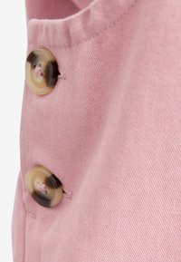 Rosa Stoff mit einer strukturierten Oberfläche, der auf der linken Seite zwei große cremefarbene und braune Knöpfe aufweist, die mit kontrastierenden Nähten befestigt sind.
