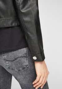 Chaqueta de cuero negra con una textura suave, con puños abotonados, combinada con unos vaqueros de mezclilla gris oscuro. Enfoque en el primer plano de la manga y la mano.