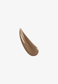 Produit crème brun avec une texture lisse et brillante, dotée d'un effet nacré, en forme de goutte courbée sur fond blanc.