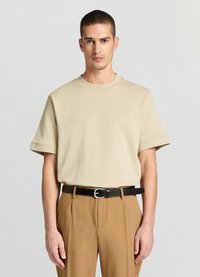 T-shirt en coton beige avec un col rond et des manches courtes, associé à un pantalon marron sur mesure et une ceinture en cuir noire.