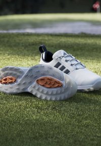 Chaussures de golf blanches avec des rayures noires sur l'herbe verte près d'un bunker de sable et d'un drapeau de trou de golf éloigné.