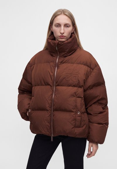 Femme portant une doudoune marron oversize à col montant, mains dans les poches, pantalon noir, debout sur un fond clair uni.