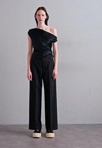 Černý top na jedno rameno z atlasu s draped materiálem, spárovaný s černými kalhotami s vysokým pasem. Zvýšené platformové boty v kontrastních barvách.