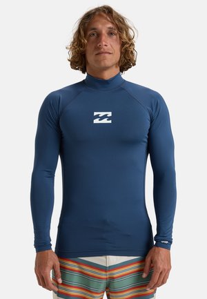 Granatowy rash guard z długim rękawem wykonany z elastycznego, odprowadzającego wilgoć materiału z wysokim kołnierzem i białym logo fali na klatce piersiowej.
