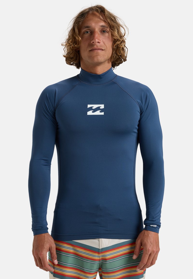 Marinblå långärmad rash guard gjord av stretchigt, fukttransporterande material med hög krage och vit våglogga på bröstet.