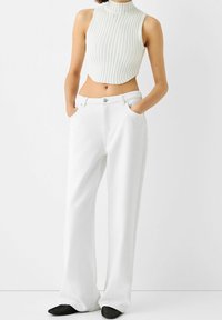 Vita högmidjade vida jeans och en ribbad, kortärmad, ljusfärgad topp utan ärmar. Modellens händer är i fickorna, skorna är svarta.